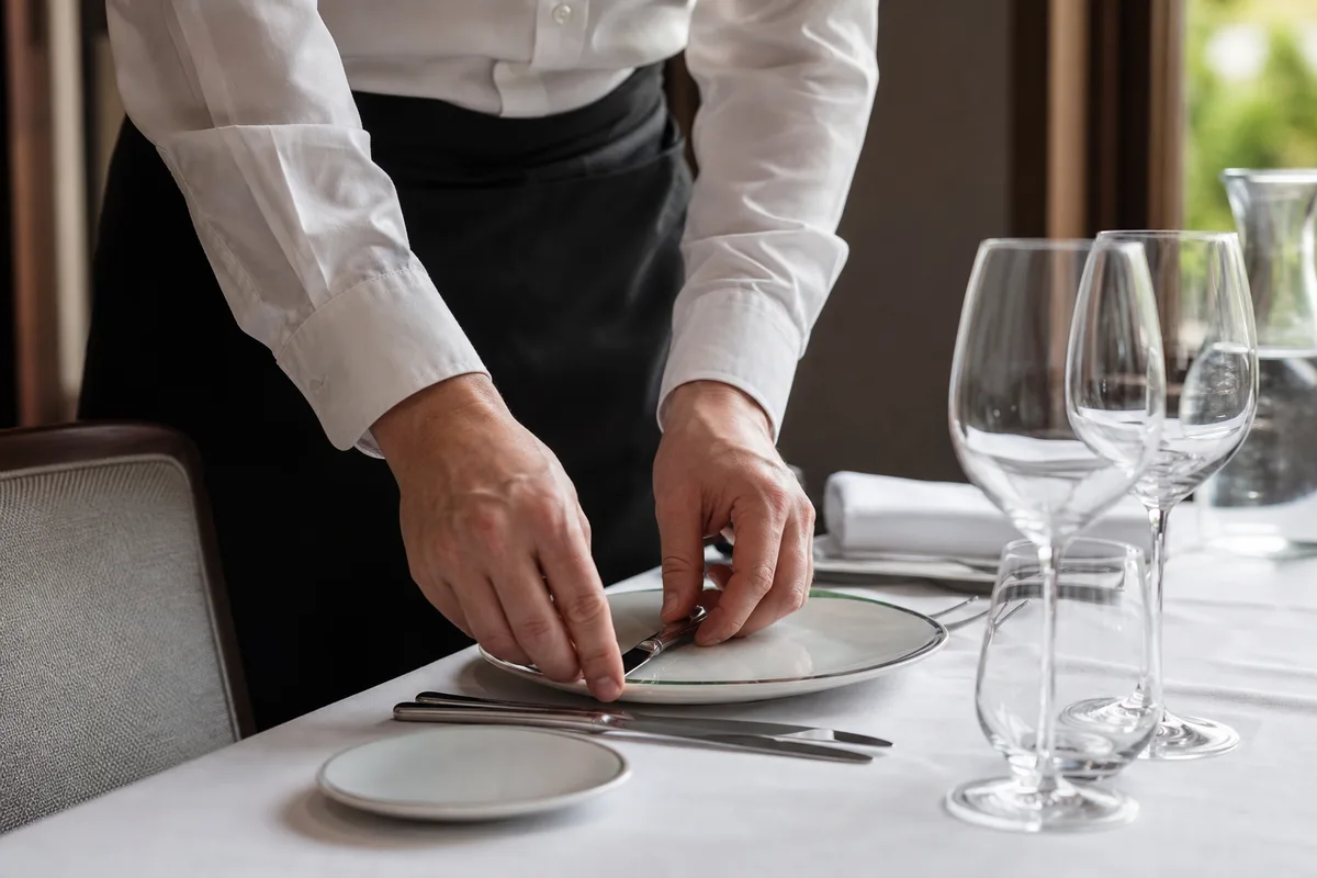 Mains dressant une table de restaurant, couverts et verres alignés sur une nappe blanche